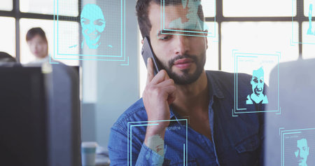 Talking on smartphone Middle Eastern man viewing monitors on office desk, with recognition overlays....