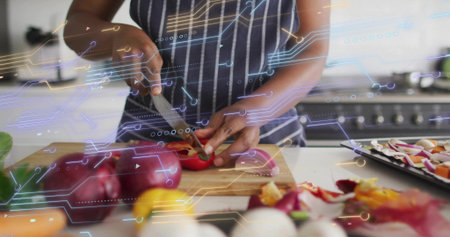 Woman wearing striped apron chopping red bell pepper on cutting board in kitchen, with chef's knife....