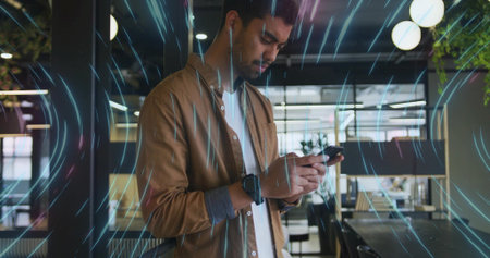 Tapping smartphone South Asian professional standing in office, with smartwatch and light streaks. Technology, innovation, workspace, connectivity, modern, professional, freelanceの写真素材