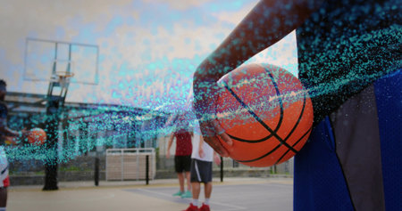 Athletic male player holding orange basketball and standing on urban court, with hoop and teammates. Athleticism, teamwork, sport, outdoor, urban, competition, activeの写真素材