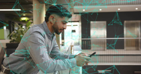 Leaning Indian man wearing gray shirt checking smartphone in office atrium with railing, backpack....