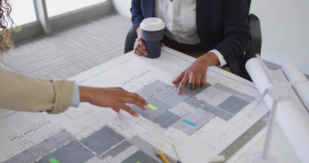 Leaning women in blazers reviewing blueprints with sticky tabs in meeting room, with coffee cup. Collaboration, planning, workspace, modern, design, teamwork, blueprintの写真素材