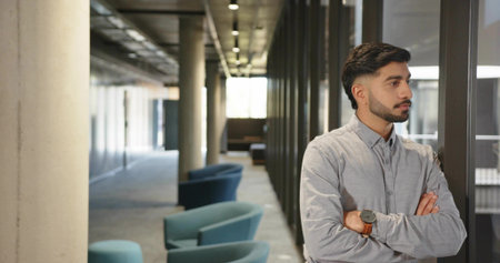 Standing Indian office worker crossing arms in hallway, with glass wall and teal chairs, copy space. Modern, professional, minimalist, corporate, architecture, workspace, innovationの写真素材