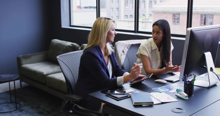 Discussing project two businesswomen collaborating at office desk, with screen tablet and notebook....