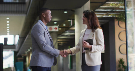 Two business professionals in blazers shaking hands in modern office corridor, with tablet. Corporate, collaboration, modern, professionalism, networking, architecture, communicationの写真素材