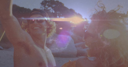 Laughing friends dancing on shoreline at beach sunset, with baseball cap and sunglasses. Joy, leisure, outdoor, vibrant, celebration, companionship, vacationの写真素材