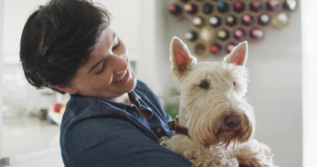 Holding small terrier dog, woman wearing dark denim shirt smiling down in kitchen, with wine rack....