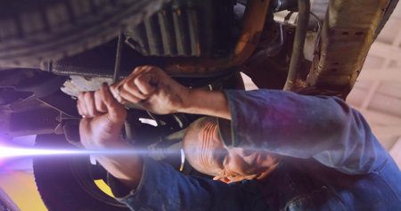 Mechanic in blue coverall tightening oil pan bolts under car lift in garage, with socket wrench. Industrial, automotive, mechanic, repair, workshop, tool, engineeringの写真素材