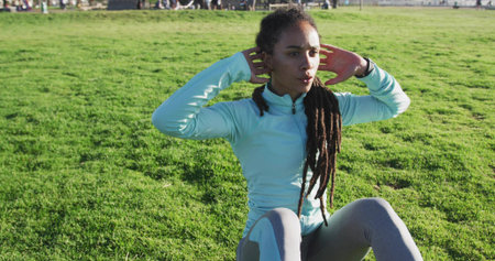 Woman in light blue jacket and gray leggings completing sit-ups on park grass, with fitness tracker. Outdoor, fitness, athleticwear, health, wellness, motivation, recreationalの写真素材