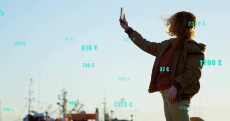 Red-haired woman holding smartphone on dock at fishing harbor, with currency overlays, copy space....