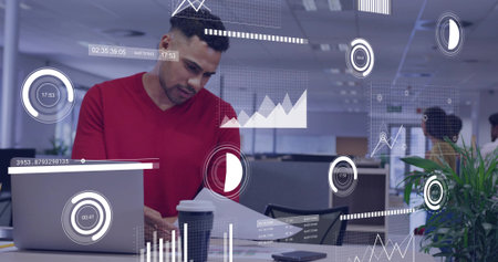 Reviewing printed charts, man in red sweater working at modern office desk, with laptop, coffee cup. Business, collaboration, technology, productivity, innovation, corporate, teamworkの写真素材