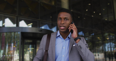 Businessman in suit talking on smartphone at building entrance, with brown shoulder bag, smartwatch....