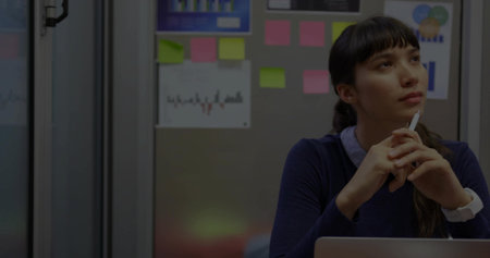 Holding pen, woman in navy sweater gazing upward in office booth with open laptop, copy space....