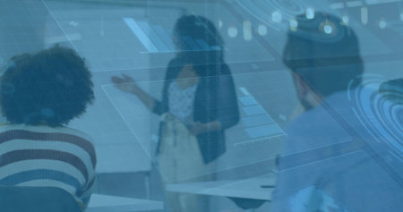 Female presenter gesturing to projected charts in meeting room with colleagues and desks....