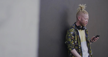 Leaning male checking smartphone at city alley with floral shirt and headphones, copy space. Urban, modern, fashionable, technology, solo, contemporary, stylishの写真素材