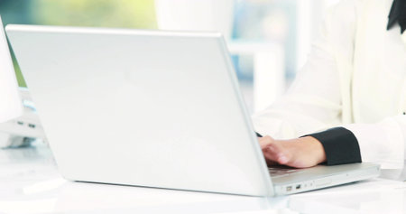 Typing woman wearing white black-cuffed blouse at desk near window using silver laptop, copy space. Professional, minimalist, modern, workspace, productivity, clean, natural lightの写真素材