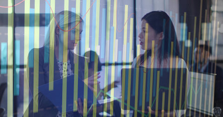 Discussing businesswomen in blazers holding blue folder, laptop in open office, with chart overlay. Collaboration, office, modern, technology, teamwork, analytics, professionalの写真素材