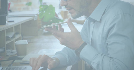 Man wearing blue shirt speaking into phone, typing on laptop in office, with coffee cup, notebook....