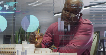 Man wearing glasses adding yellow pencil notes to scale model in design studio, with digital charts....