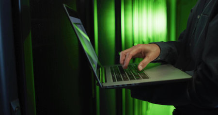 Technician typing commands on laptop by server rack, with network cables, LEDs, copy space....