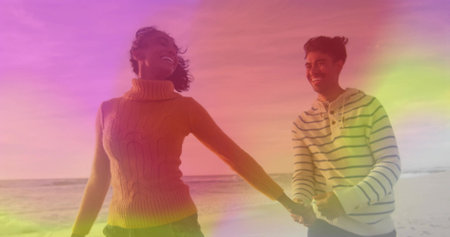 Walking couple wearing mustard cable-knit turtleneck and striped hoodie on sandy shoreline, smiling. Romance, connection, outdoors, leisure, serenity, happiness, casualの写真素材