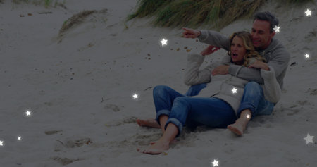 Pointing mature couple wearing jeans and sweaters cuddling on sandy dunes at beach, copy space....
