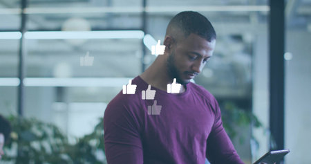 Focusing man wearing maroon t-shirt reviewing tablet in open office, with floating thumbs-up icons. Technology, engagement, modernity, social media, communication, digital, corporateの写真素材