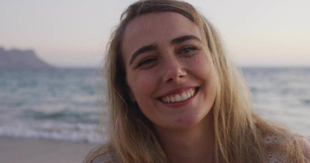 Smiling adult female wearing sleeveless knit top standing on sandy beach at dusk, soft sunset light. Serenity, leisure, outdoor, natural, tranquil, relaxed, beautyの写真素材