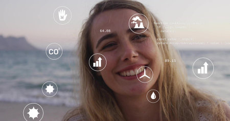 Smiling blonde woman standing on beach with digital overlays showing CO2 icon, other icons and code. Environment, sustainability, innovation, technology, wellness, advocacy, natureの写真素材