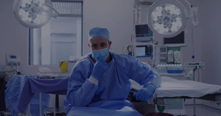 Sitting surgeon wearing surgical attire observing monitors in operating room, with overhead lights....