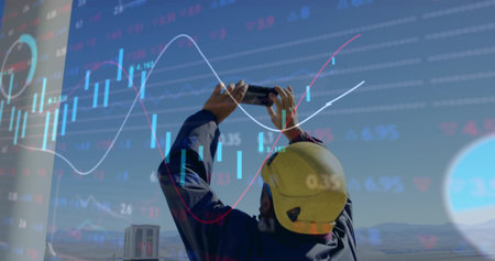 Field worker in coveralls and helmet raising smartphone at site, with financial chart overlay....