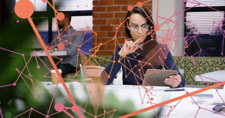 Asian woman reviewing tablet while holding pencil to lips in modern office, with potted plant. Collaboration, teamwork, modern, technology, productivity, business, connectivityの写真素材