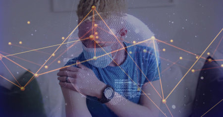 Man wearing blue t-shirt and medical face mask holding left shoulder on couch, with network overlay. Health, technology, connectivity, telemedicine, innovation, digital, wellnessの写真素材