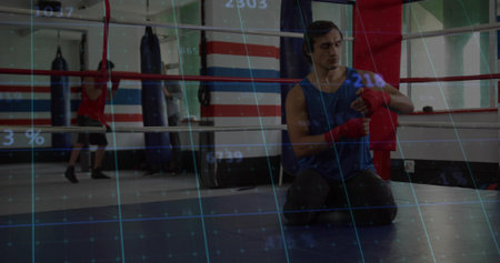 Male boxer wrapping red hand wraps around wrists in boxing ring area, with punching bags. Athlete,...