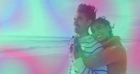 Embracing couple wearing tan and striped sweaters gazing toward calm sea on sandy beach, copy space....