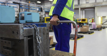 Warehouse worker in safety vest pushing hand truck in factory hall, with mold block and chains. Industrial, logistics, construction, equipment, safety, manufacturing, efficiencyの写真素材