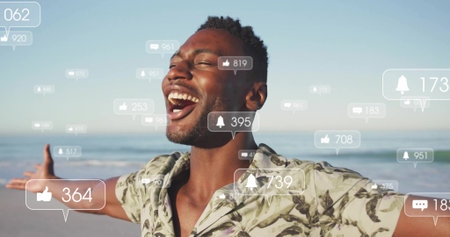 Laughing male wearing tropical shirt standing on sandy shore, with floating notification icons. Joy, relaxation, celebration, coastal, vibrant, casual, digital communicationの写真素材