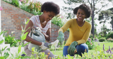 Kneeling mother and daughter pouring water on flowering plants in backyard, with gray watering can. Gardening, nurturing, family, flora, outdoor, harmony, growthの写真素材