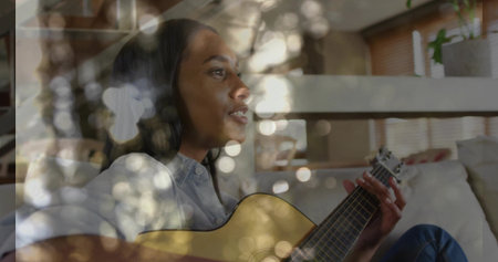 Strumming acoustic guitar musician wearing denim outfit on couch in living room, with bokeh lights....