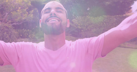 Smiling man wearing pink short-sleeved shirt tilting head back on sunlit park lawn with lens flares....