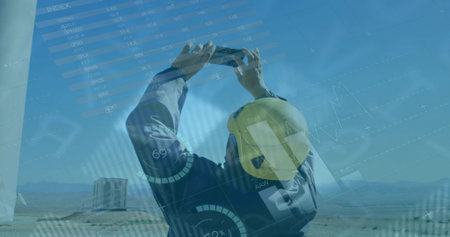 Inspecting technician wearing helmet and holding tablet near turbine tower on wind farm, with data....