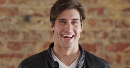 Smiling mid adult male wearing leather jacket posing against brick wall, with warm lighting. Portrait, confidence, casual, urban, contemporary, professionalism, warmの写真素材