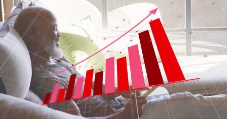 Holding tablet, senior man analyzing digital bar chart on living room sofa with green throw pillows. Retirement, relaxation, digital, analytics, comfort, home, modernの写真素材