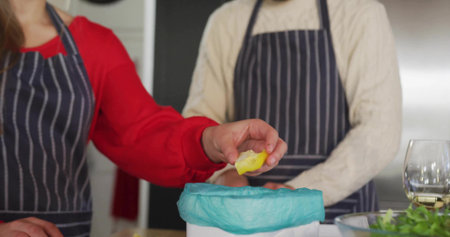 Couple wearing aprons holding lemon peel, disposing into blue bin on kitchen counter, wine salad. Domestic, culinary, rustic, cheerful, teamwork, cozy, lifestyleの写真素材