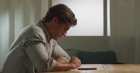 Leaning forward man in grey shirt inspecting smartphone on wooden table in living room, copy space....
