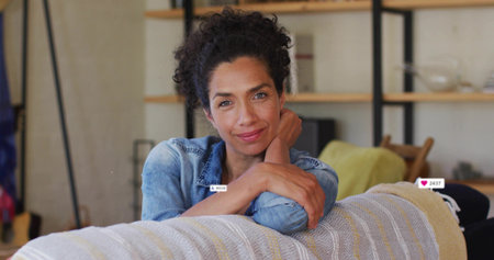 Leaning woman resting arms on sofa in living room, wearing denim shirt with metal shelving unit....
