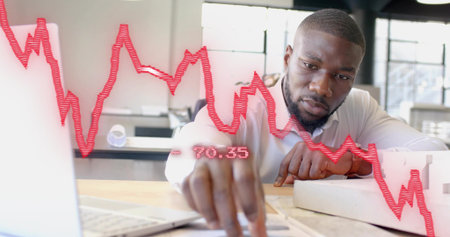 Reviewing documents on laptop, African American man sitting in modern office with red chart overlay....
