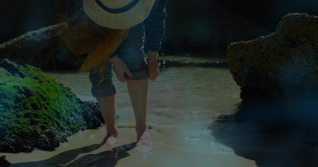 Bending woman rolling up denim pant cuffs at seaside tide pool, with straw hat, sunlit water. Nature, outdoor, leisure, adventure, exploration, tranquility, sunlightの写真素材