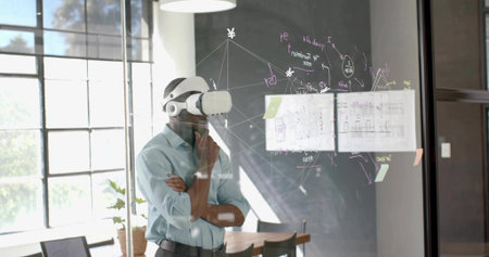 Gazing man wearing business attire and VR headset at glass partition with diagrams in meeting room....