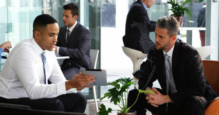 Discussing data on tablet two professionals wearing suits in lounge with potted plants on tables. Modern, collaboration, interior, workspace, greenery, technology, meetingの写真素材